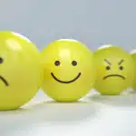 Four yellow spherical emojis or balls with different facial expressions lined up from left to right: a sad face, a happy face, an angry face with furrowed brows, and a worried or concerned face with tears. They are on a light grey surface.