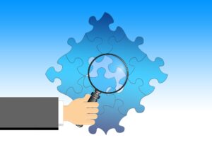A hand holding a magnifying glass over a blue puzzle, with one piece highlighted, illustrating the concept of breaking down complex tasks and focusing on smaller parts for learning.