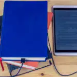 A stack of physical books (blue and orange) with a charging cable plugged into the bottom book, leading to a tablet displaying text.