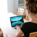 A woman with red hair in a bun, seen from behind, working on a laptop at a light-colored wooden table in a home setting.