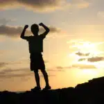 A silhouette of a child with arms raised in triumph against a vibrant sunset sky, symbolizing the victory and progress achieved by children in speech therapy.