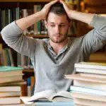 Stock photo of a man sitting at a library table piled high with books, with his hands in his hair, looking overwhelmed, symbolizing the stress of large academic tasks or continuing education.
