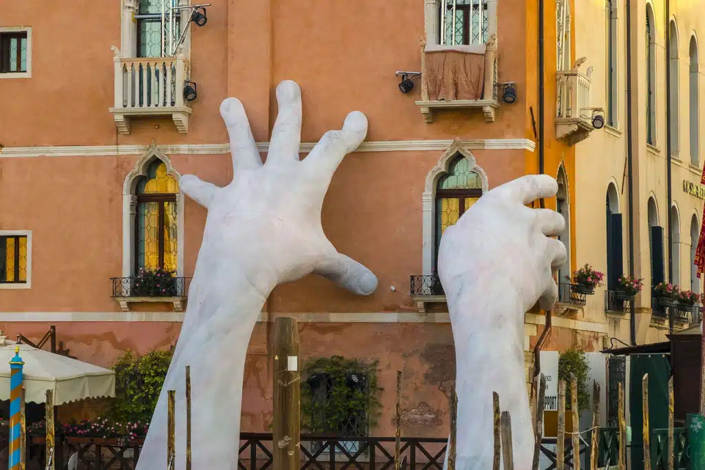 Lorenzo Quinn's "Support" sculpture in Venice, featuring two monumental white hands emerging from the Grand Canal and reaching towards a building.