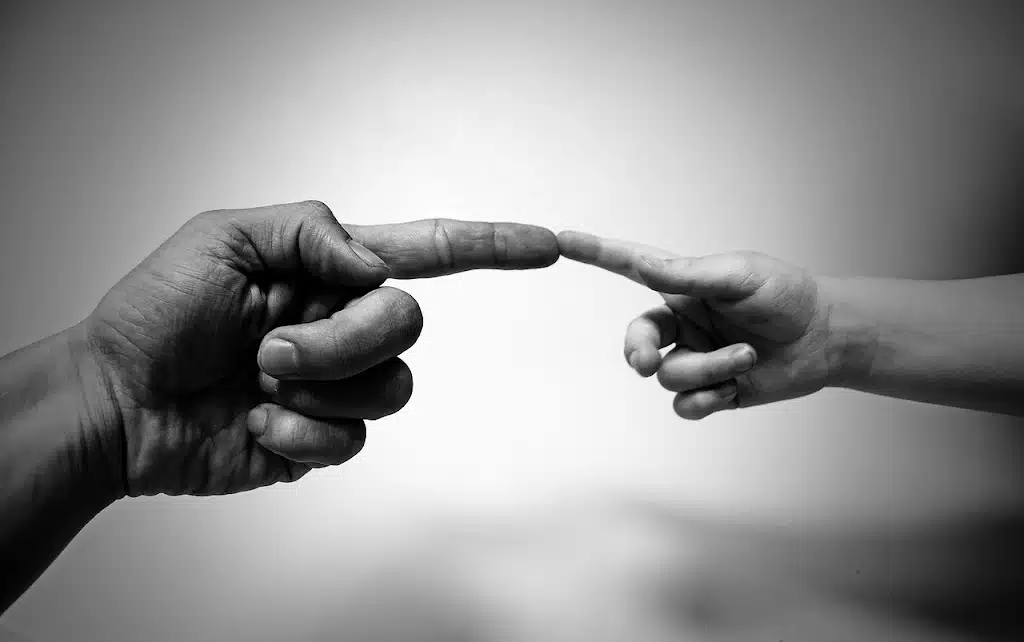 A black and white close-up image showing a larger adult hand and a smaller child's hand reaching towards each other, with their index fingertips almost touching in the center, symbolizing connection or guidance.
