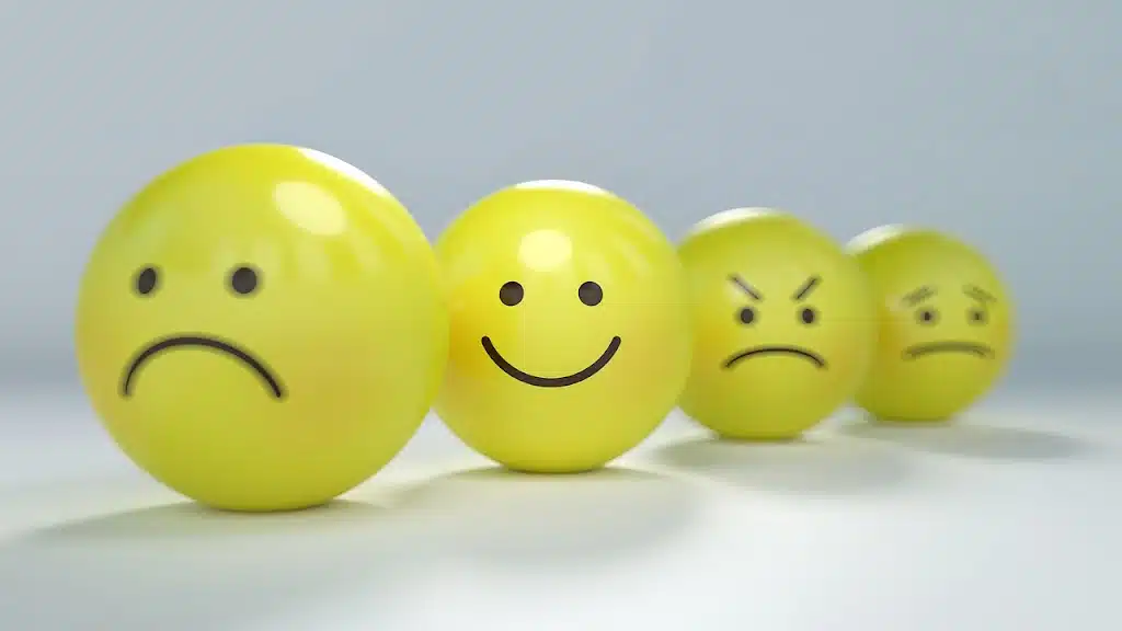 Four yellow spherical emojis or balls with different facial expressions lined up from left to right: a sad face, a happy face, an angry face with furrowed brows, and a worried or concerned face with tears. They are on a light grey surface.