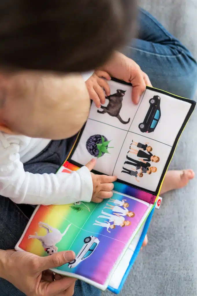 Photograph of an adult and an infant sitting together, with the infant actively engaged and touching colorful images in a baby book, symbolizing early language development through pictures.