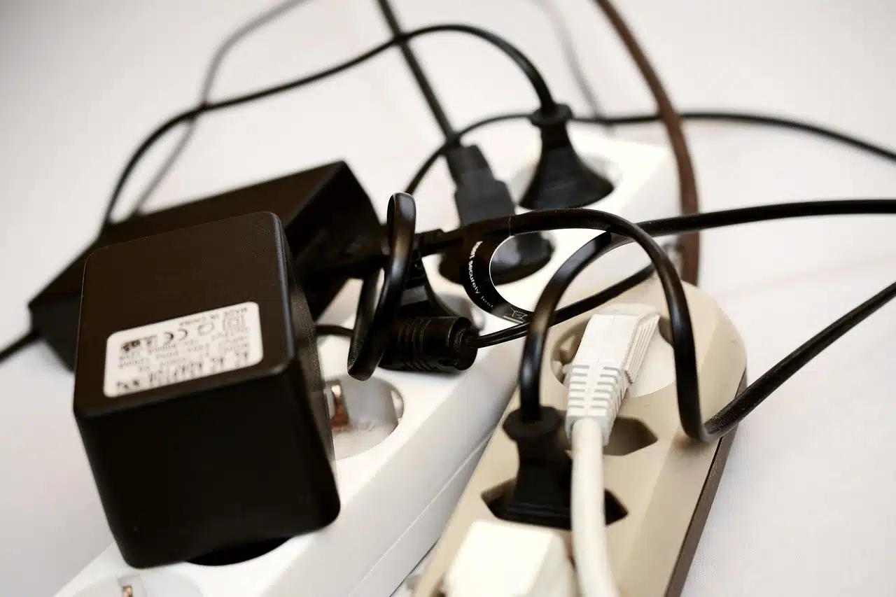 A close-up of multiple black power adapters and cables densely plugged into white and beige power strips, creating a tangled and busy appearance.
