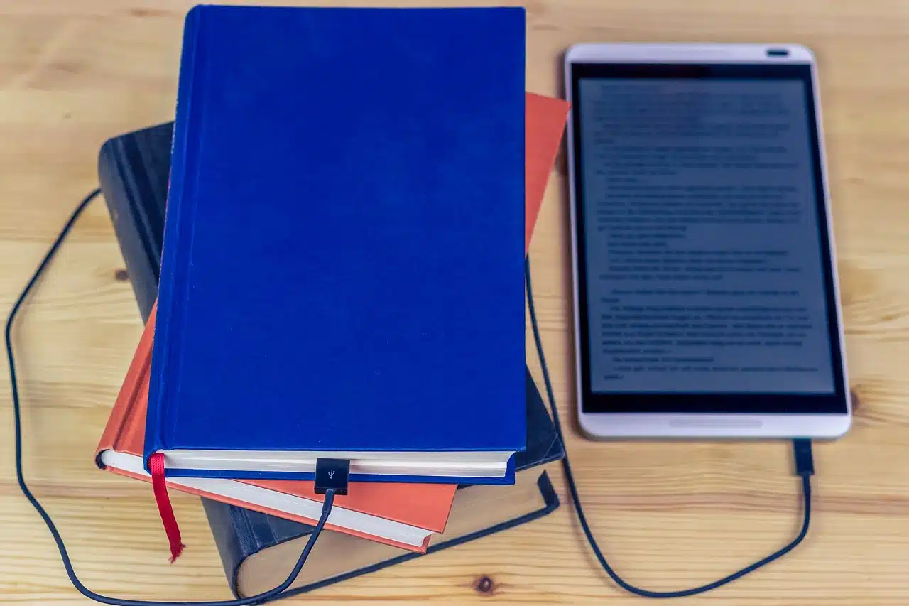 A stack of physical books (blue and orange) with a charging cable plugged into the bottom book, leading to a tablet displaying text.