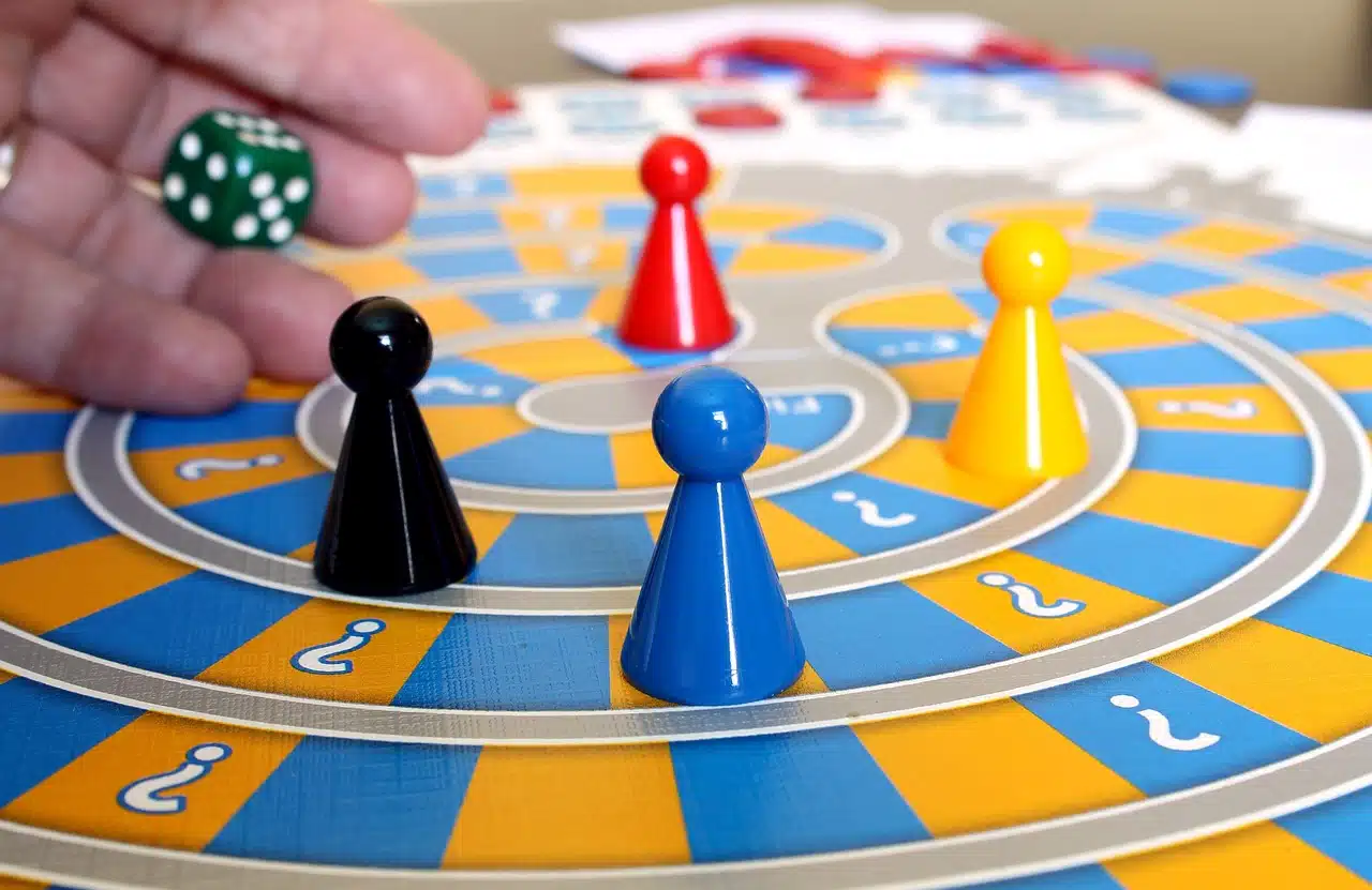 Hand rolling a green dice over a colorful board game with black, red, yellow, and blue pawns, representing gamification in speech therapy.
