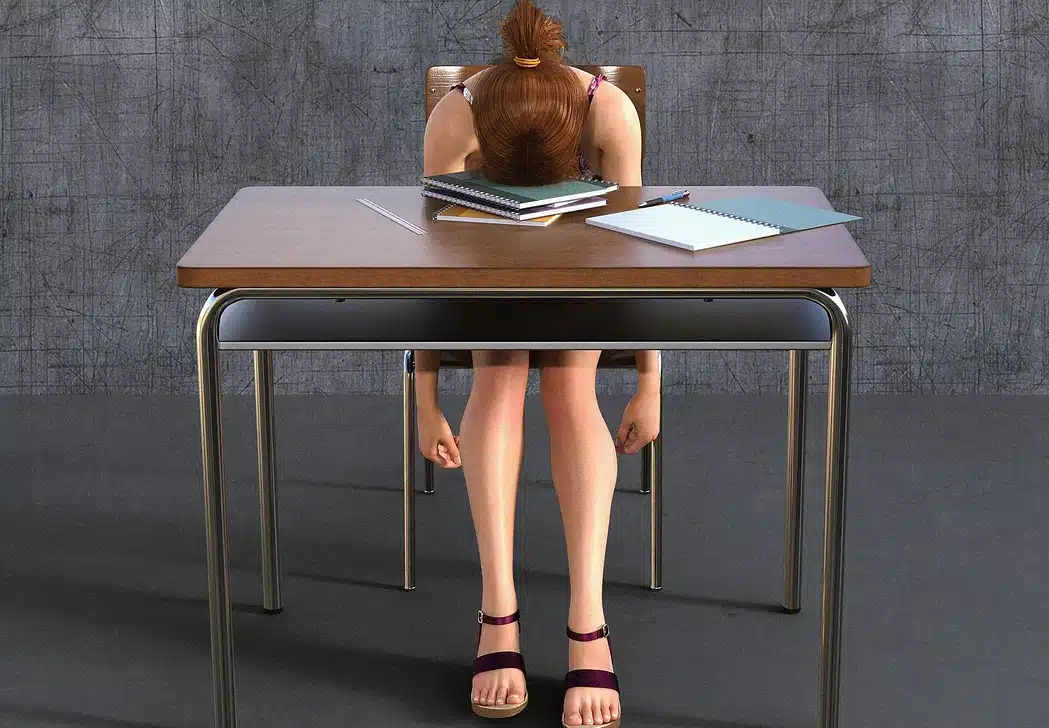 A student with their head down on a school desk, surrounded by notebooks and a pencil, appearing tired, bored, or overwhelmed. Only the student's back and legs are fully visible.