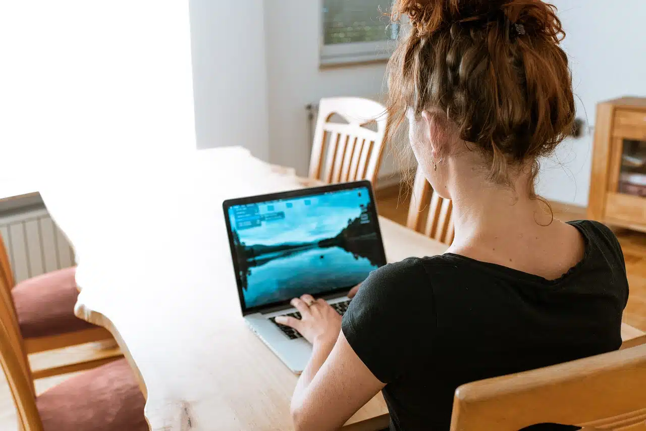 A woman with red hair in a bun, seen from behind, working on a laptop at a light-colored wooden table in a home setting.