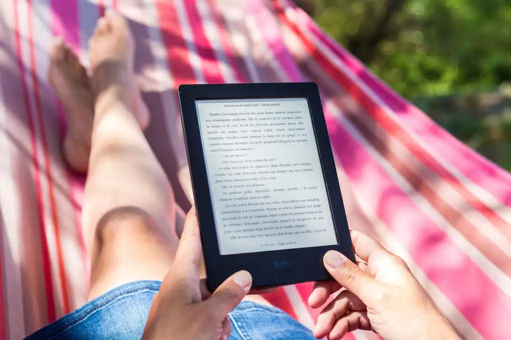 A person relaxing in a red and white striped hammock, holding a black e-reader displaying text. Their bare feet are visible in the foreground.