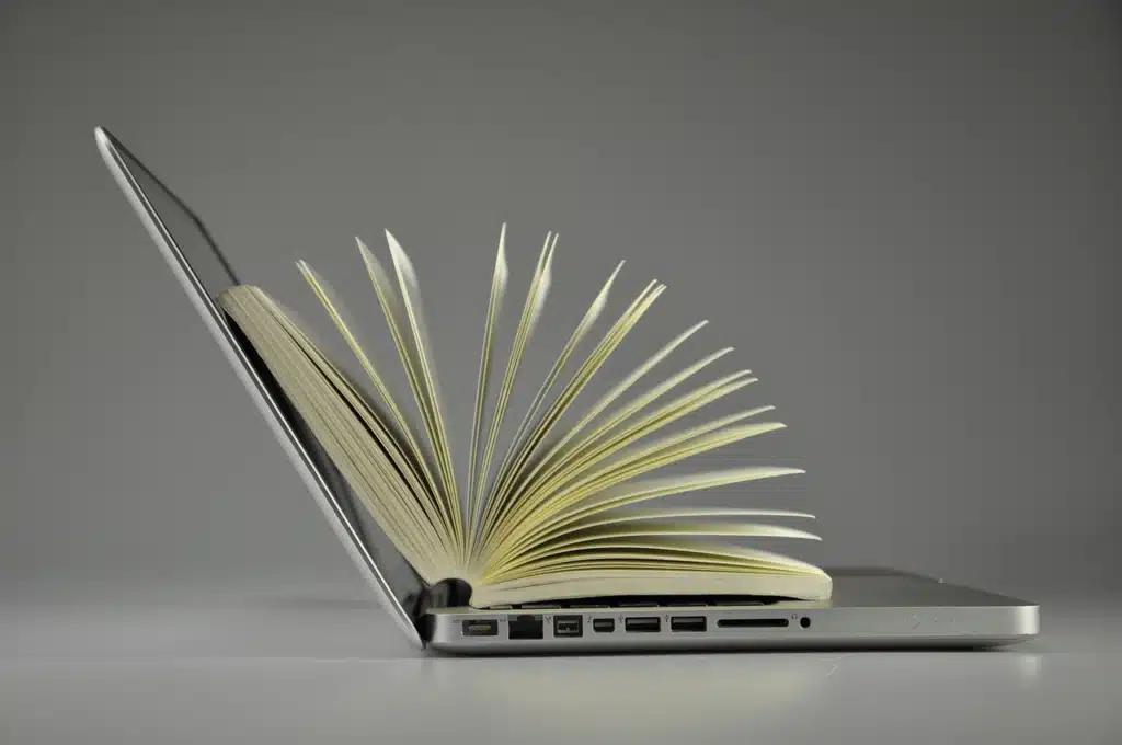 An open physical book with pages fanned out, resting on the keyboard of an open silver laptop.