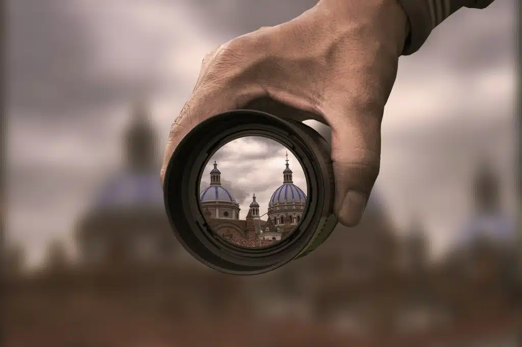 A person's hand holding a camera lens in the foreground, through which distant domed buildings are seen clearly in focus, while the immediate background around the lens is blurred.