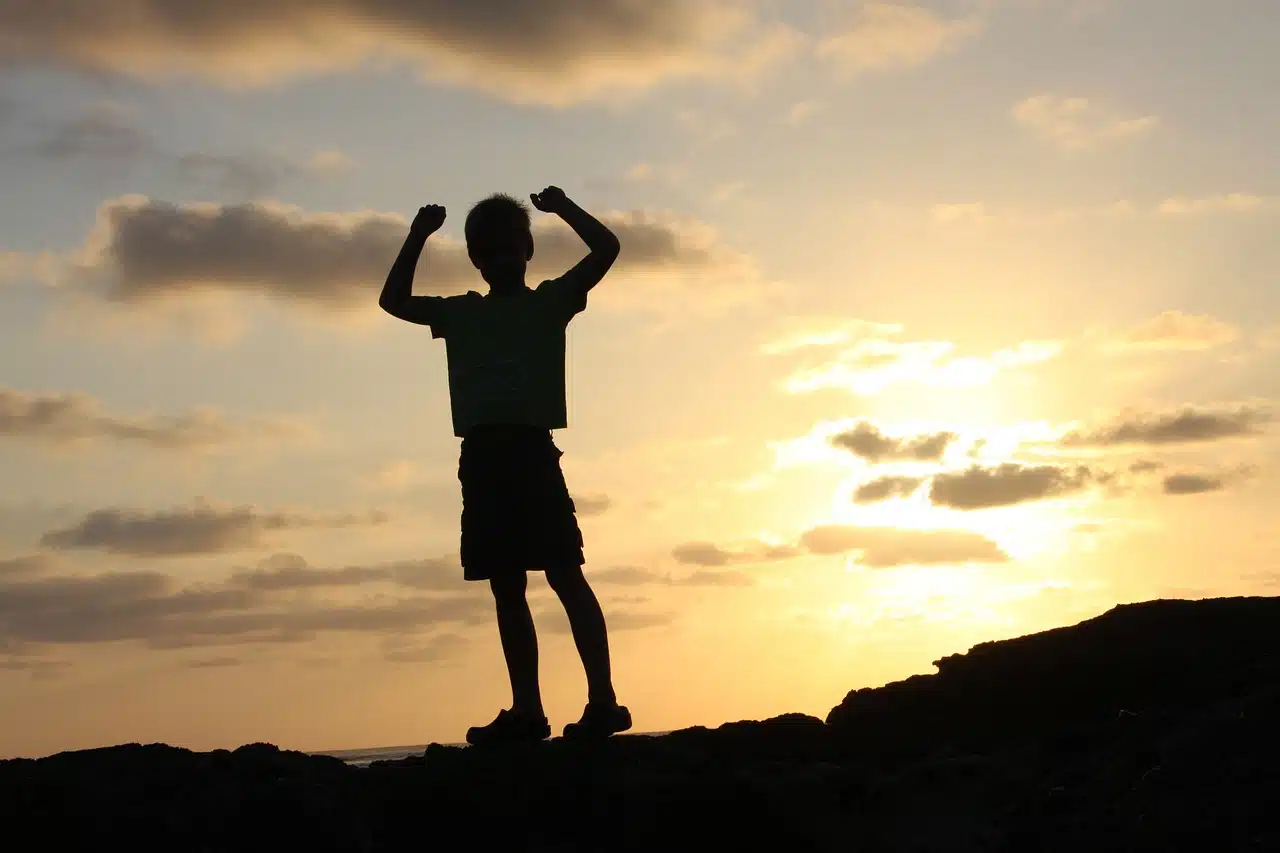 A silhouette of a child with arms raised in triumph against a vibrant sunset sky, symbolizing the victory and progress achieved by children in speech therapy.