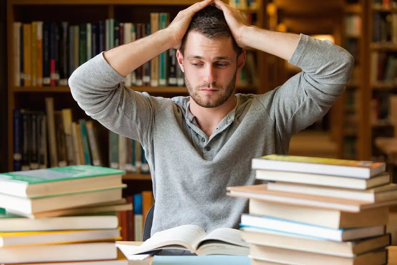 Stock photo of a man sitting at a library table piled high with books, with his hands in his hair, looking overwhelmed, symbolizing the stress of large academic tasks or continuing education.