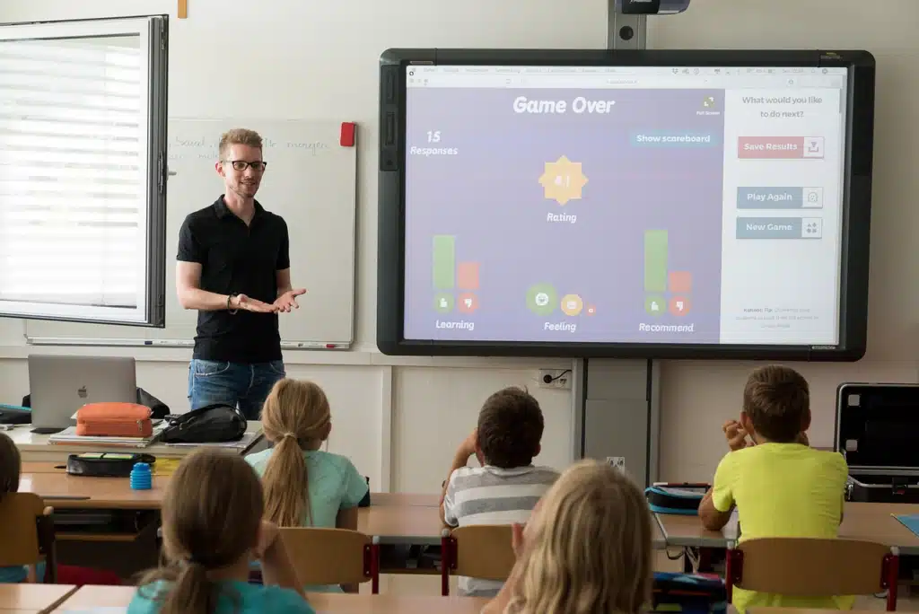 A male teacher with glasses smiles as he gestures towards a large interactive smart board displaying a game, engaging a classroom of elementary students, symbolizing a positive and stimulating learning environment.