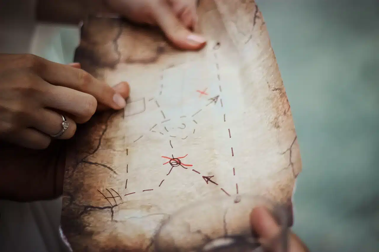 Two pairs of hands holding a weathered treasure map with drawn lines, dotted paths, an arrow, and a circled "X" marking a spot. One finger points towards the marked location.