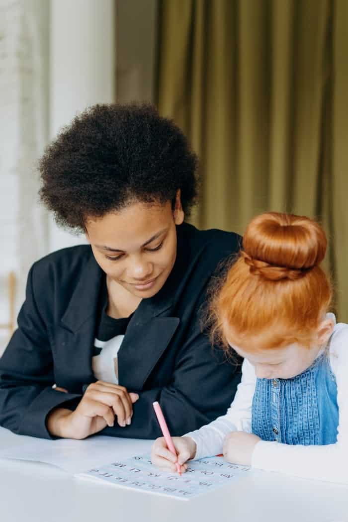 about-img A teacher assists a student with her homework at a desk indoors, promoting learning and education.