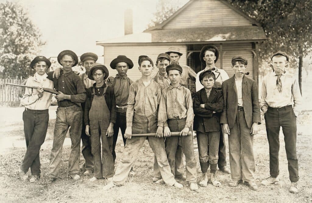 Sepia photo of a group of rough-looking older boys in early 1900s work attire posing defiantly outdoors near a building, with two boys holding a rifle.